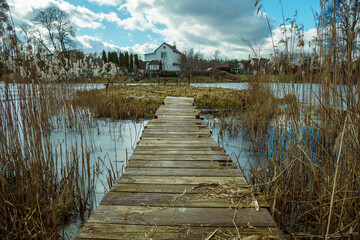 A wooden bridge leads from the shore to a small island on the lake. Fishing island on the background of clouds. A village and private houses on the shore of a clear lake.