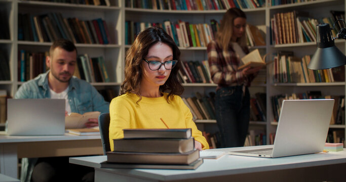Young female student in glasses studying n library at laptop computer and getting bored. Boring study in bibliotheca concept. Girl learning and preparing to exam.