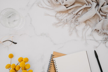 Flat lay of white notebook with copy space for text, glasses, glass of water and yellow flowers on white marble background