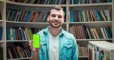 Portrait of young Caucasian handsome male student smiling and showing smartphone with green screen...