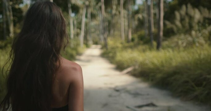 Young hiker woman with long brown hair, back view, walking in jungle forest. Girl hiking alone. Tourist on jungle park trail, looking around. Exotic travel, tourism, holiday, hiking, active lifestyle.