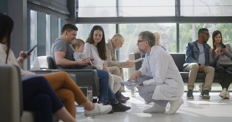 Diverse people sit on couches in clinic lobby area, wait for doctors appointment. Doctor talks to...