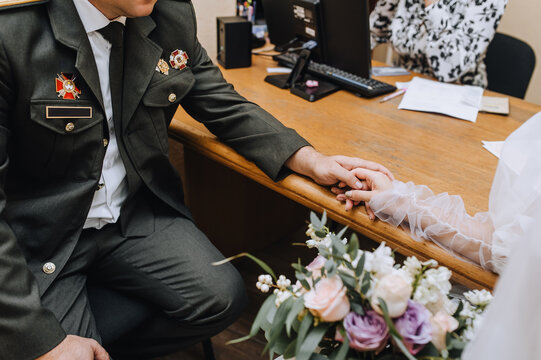A military man soldier, a groom in uniform and a beautiful bride in a white dress are sitting in the registry office holding hands at the ceremony. Wedding photography, portrait.
