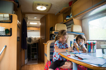 Two caucasian children, brother and sister, drawing inside a campervan during a road trip stop. Camper vacations travelling with kids background.