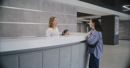 Adult woman stands at reception desk in clinic lobby entrance and makes appointment with doctor. Female administrator talks with patient at information counter. Medical staff work in modern hospital.