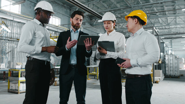 Teamwork at background of working process where welding sparks flying. Multicultural industrial engineers in hard hats use tablet and have discussion at construction site. Ethnic diversity.