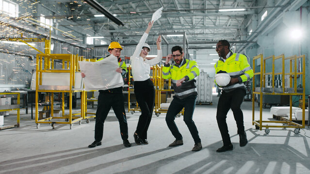 Multicultural engineers dancing at modern manufacturing factory and clapping hands. Employees in safety hard hats celebrating succsess at background of welding sparks flying.
