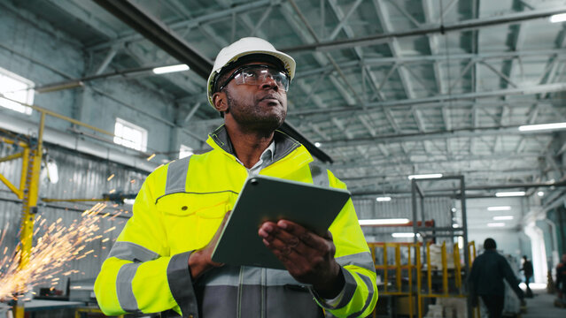 African American engineer in hard hat and protective vest is looking through workshop. Man is holding gadget and gesturing at construction site. Employee on background of welding sparks flying.