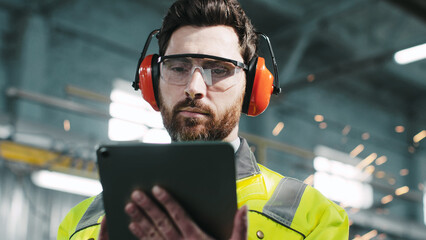 Bearded worker in protective headphones and googles wearing reflective yellow vest is looking at...
