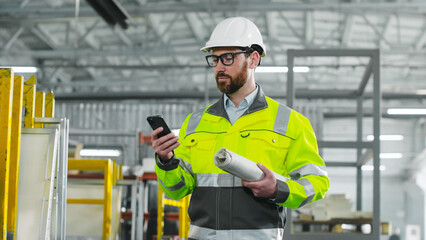 Main engineer monitors work process. Man in protective glasses and reflective clothes is holding phone. Worker with blueprint. Male is going at workshop on background of workflow and equipment.