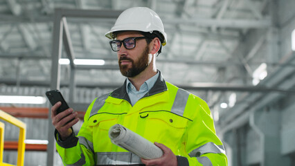 Bearded architect inspect ndustrial site. Male holds blueprint and look at phonescreen. Industrial man in reflective vest and hard hat at construction site. Worker at background of work process.