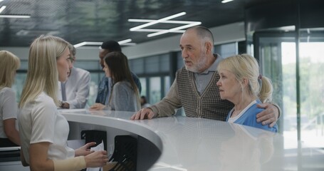 Obraz premium Elderly couple stand near reception desk in clinic lobby area, come on appointment with doctor. Female receptionist talks with patients at information counter. Medical staff work in modern hospital.