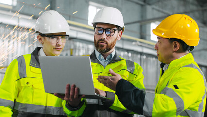 Three construction workers in half height look at laptop. Engineers in uniform collaborate with each other in workshop. Man in yellow helmet explains plan of action to employees.