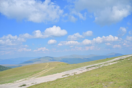 Stunning Summer Aerial View: Glorious Light Clouds in a Crisp Blue Sky Over Mountain Peaks with a Scenic Land Path Leading from the Summit
