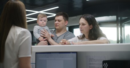 Family with little child stands near reception desk in clinic lobby area. Woman fills out documents and papers, makes appointment with doctor. Medical staff work in modern medical center. Health care.