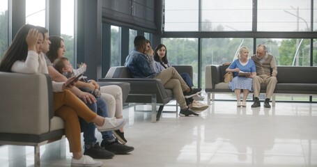 Diverse multiethnic people sit on couches in clinic lobby area, wait for appointment with doctor or...