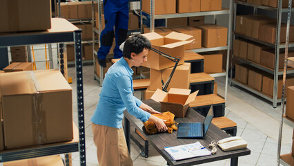 Woman employee taking supplies from warehouse shelves and putting them in cardboard boxes, using packages for delivery. Young adult writing inventory information on laptop, logistics.
