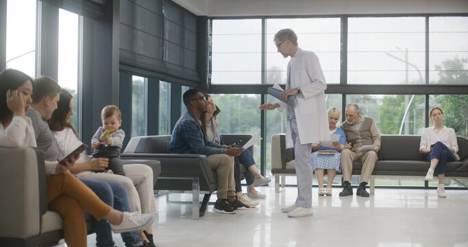 Diverse people sit on sofas in clinic lobby area, wait for appointment with doctor. Doctor speaks with multiethnic couple about medical test results. Waiting area in modern medical center. Healthcare.