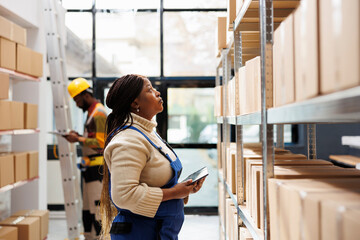 Logistics and distribution manager inspecting merchandise storage in warehouse. African american storehouse employee standing near parcels shelf, holding tablet and looking at carton