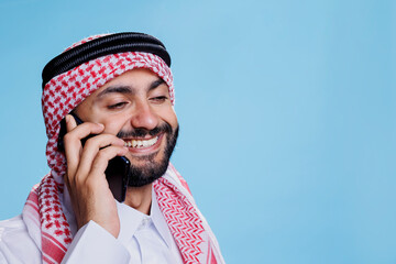 Smiling muslim man wearing ghutra headscarf with rope band talking on mobile phone. Cheerful arab dressed in islamic headdress speaking on smartphone with happy expression
