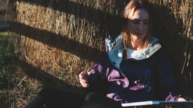 Woman with laptop relaxes in countryside sitting near a stack of hay. Caucasian girl working on notebook and enjoy nature landscape. Concept of freelance, digital remote online work or business