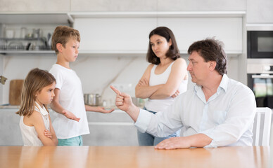 Angry parents scolding two children standing in front of them disappointedly in the kitchen