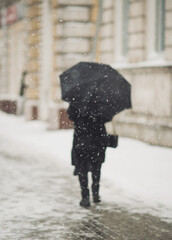 Blured figure of old woman with umbrella in warm clothes walking in the street in snowy winter day. First snow. Spending time alone in the city. Back view. figure of lonely woman going away.