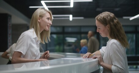 Female administrator talks with patient at reception desk in clinic lobby area. Woman consults...