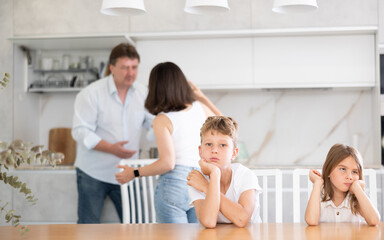 Frightened and frowning children sit at kitchen table during mom and dad quarrel. Behind children, parents are swearing in background. Family conflict, divorce