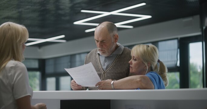 Elderly couple stand near reception desk in clinic lobby area, look at tests results on paper. Female receptionist at information counter. Medical staff work in modern hospital or medical center.