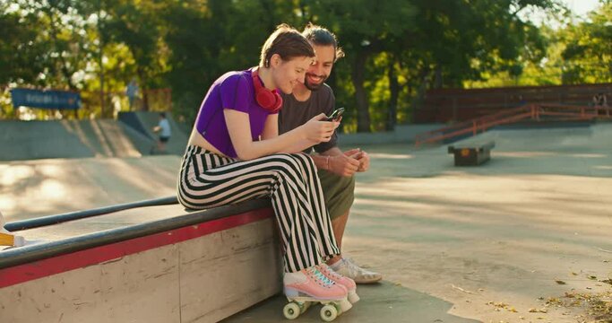 Side view of a girl with a short haircut in a purple top striped pants in pink roller skates sits on a bench in a skate park and communicates with a brunette guy in a gray t-shirt with a phone in her
