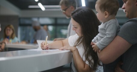 Family with little child stands near reception desk in clinic lobby area. Woman fills out documents and papers, makes appointment with doctor. Medical staff work in modern medical center. Health care.