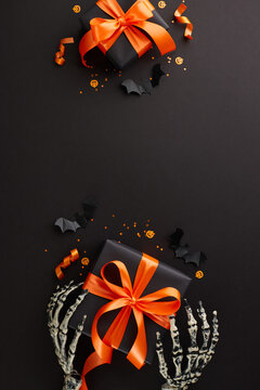 Infusing Halloween with warmth through considerate presents. Top view vertical photo of skeleton hands, dark presents, spooky bats, confetti on black background with ad placement