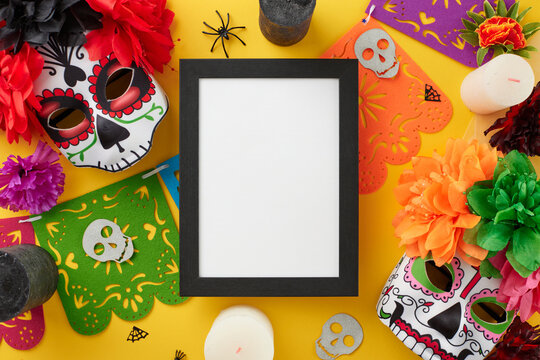 Immerse in the beauty of Dia de los Muertos traditions. Top view photo of traditional masks, colorful garland, flowers, candles, scary decor on yellow background with empty frame for ad or text