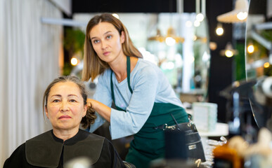 Positive elderly woman visiting professional hair salon. Skilled female hairdresser offering hairstyling to client .