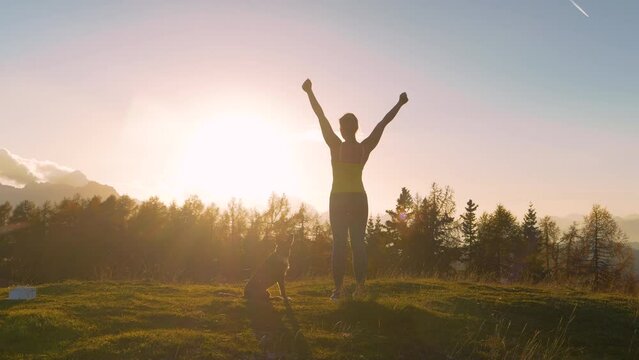 CLOSE UP, LENS FLARE: Woman victoriously raises arms on hilltop in golden light. She is celebrating reaching a picturesque peak after running uphill with her dog. Beautiful autumn day in the mountains