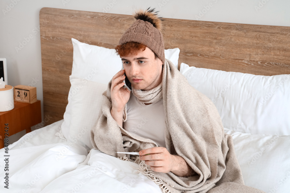 Sticker ill young man with thermometer talking by mobile phone in bedroom