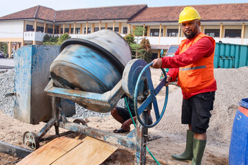 Construction worker wearing safety yellow helmet, boots and vest standing near concrete mixer at...