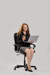 Young businesswoman with laptop and cup of coffee sitting in chair on light background