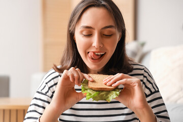 Beautiful young woman eating tasty sandwich in living room, closeup