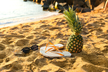 Obraz premium Stylish flips flops with pineapple and sunglasses on sand near ocean at resort