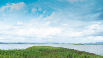 Ground covered with green grass juts out into the water. Large reservoir surrounded by mountains Under blue sky and white clouds. Pra Sae Reservoir Rayong Thailand. 