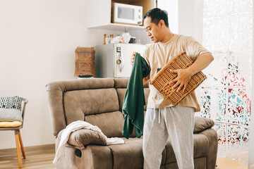 Asian man with laundry basket, picking up dirty shirts from sofa at living room