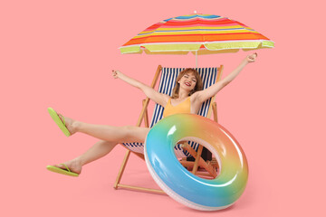 Young woman in deck chair with beach umbrella and swim ring on pink background