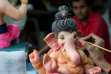 Hand of an artist painting eye of Lord Ganesha's idol in workshop with huge colourful idols for...