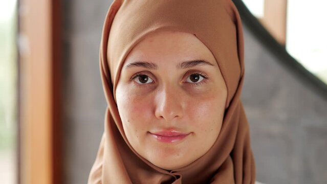 A humble young woman with her hair covered by a hijab looks down, then lifts her head and looks straight into the camera with a smile. Close portrait shot with blurred background