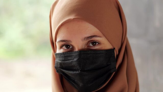 Friendly young woman with her hair covered by hijab and black medical mask on her face covering mouth and nose. She looks amiably straight into the camera and smile, welcoming and positive attitude