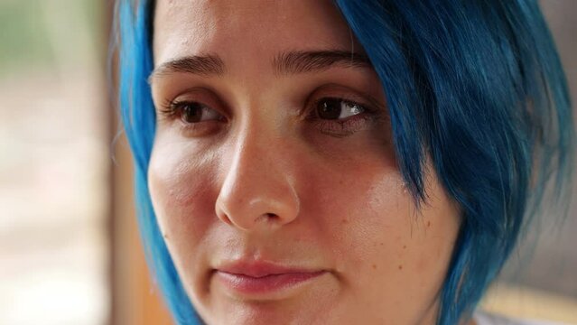 Very close portrait of face of young weeping woman, tears coming from her eyes. Woman have blue dyed hair, look aside and emotionally move lips. She feel sorrow or bad because of some news or people