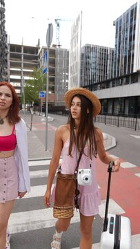 A duo of spirited young ladies is trekking through the urban streets, accompanied by a suitcase and their belongings.