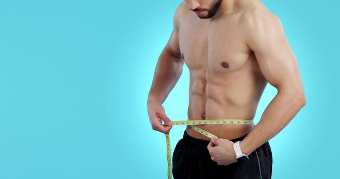 Shirtless, body and a man measuring waist in studio for weight loss, diet or gain. Athlete, bodybuilder or hands of sports person with tape on blue background for six pack and fitness progress